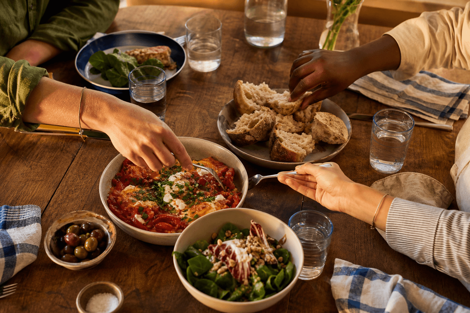 Friends' hands reaching across a wooden table for shared tomato beans, salad, olives and torn bread